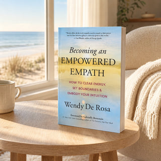 The book 'Becoming an Empowered Empath' by Wendy De Rosa is displayed upright on a light wood table in a cozy living room setting, with a window, beach view, and a cream-colored armchair with a fringed throw blanket in the background.