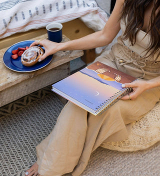 Person sitting on a bed holding a spiral-bound notebook with a plate of food and a cup nearby.
