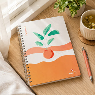 A spiral-bound notebook with a minimalist orange and white cover design featuring a potted plant illustration, styled on a wooden desk next to a pen, a small plant, and a cup of chamomile tea.