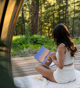 Woman sitting on a blanket in a forest, reading a book.