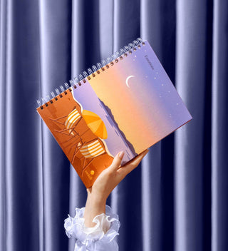 Hand holding a spiral-bound notebook with artistic cover design against a blue curtain background