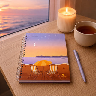 A spiral-bound notebook with a beach sunset illustration sits on a wooden table next to a lit candle, a cup of tea, and a pen. The notebook cover shows a purple sky, crescent moon, stars, ocean, and beach chairs under an orange-yellow umbrella.